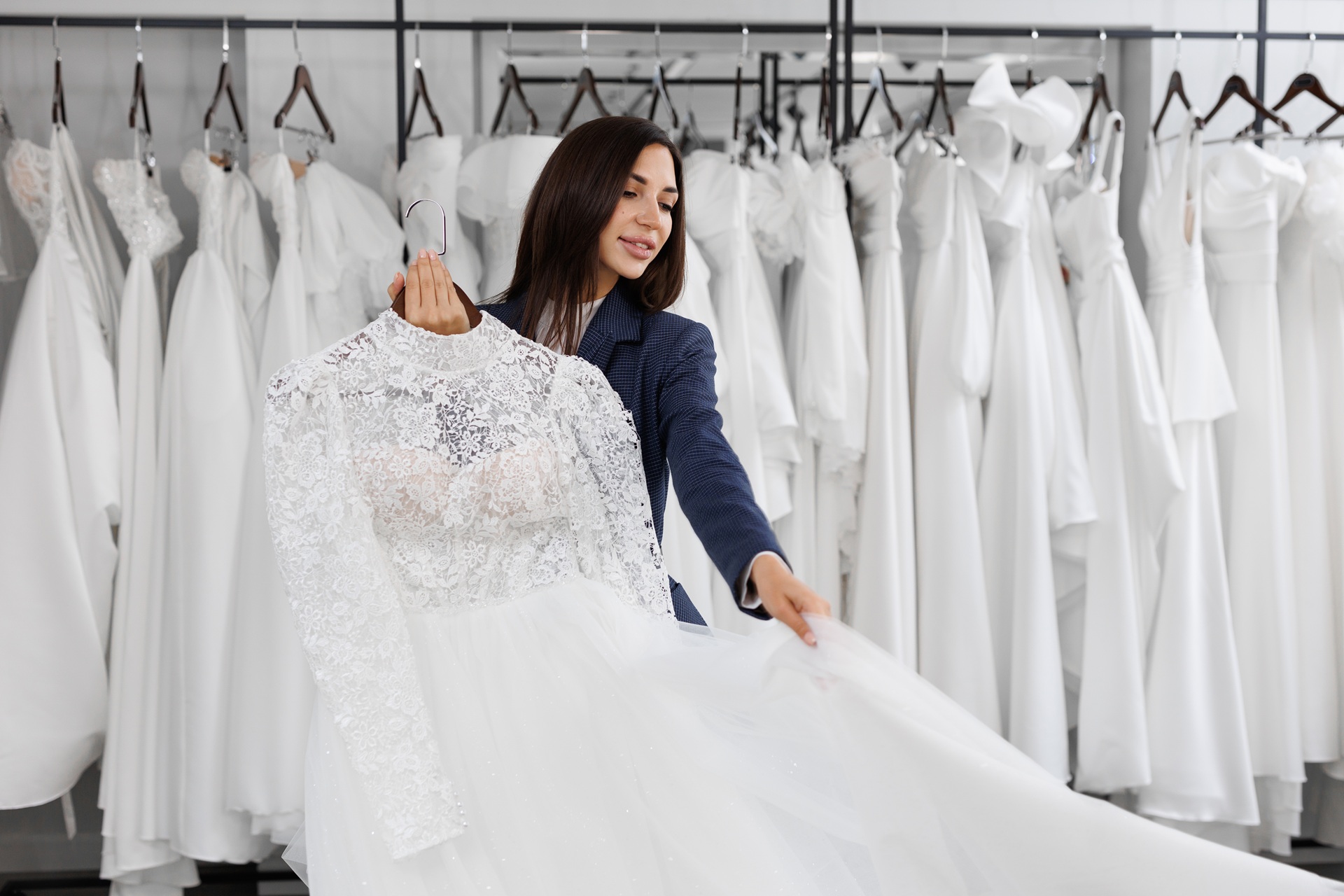 A woman in a dark suit showcases a white lace bridal gown in a store with multiple wedding dresses hanging on racks in the background.