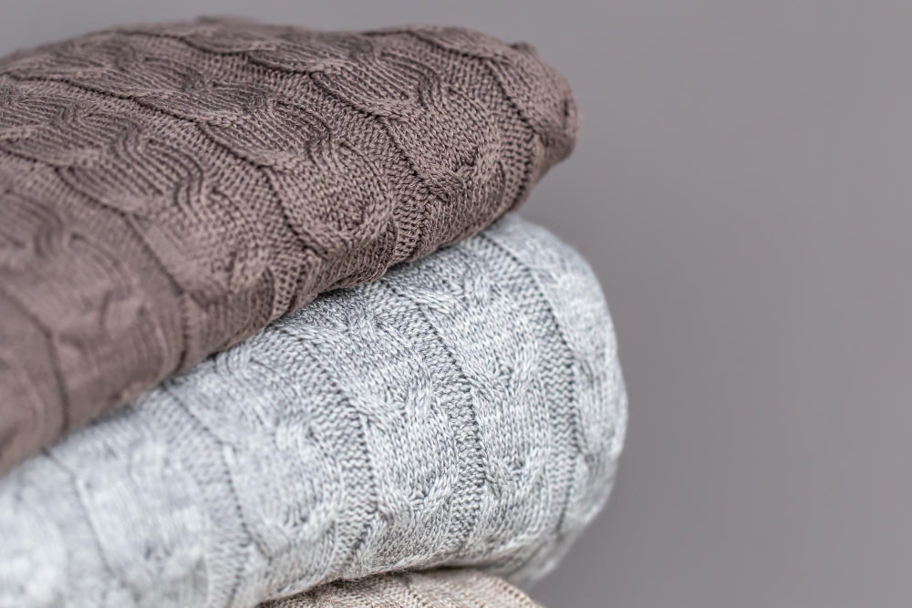 Three folded knitted sweaters in brown, gray, and beige colors are stacked on top of each other against a plain grey background.