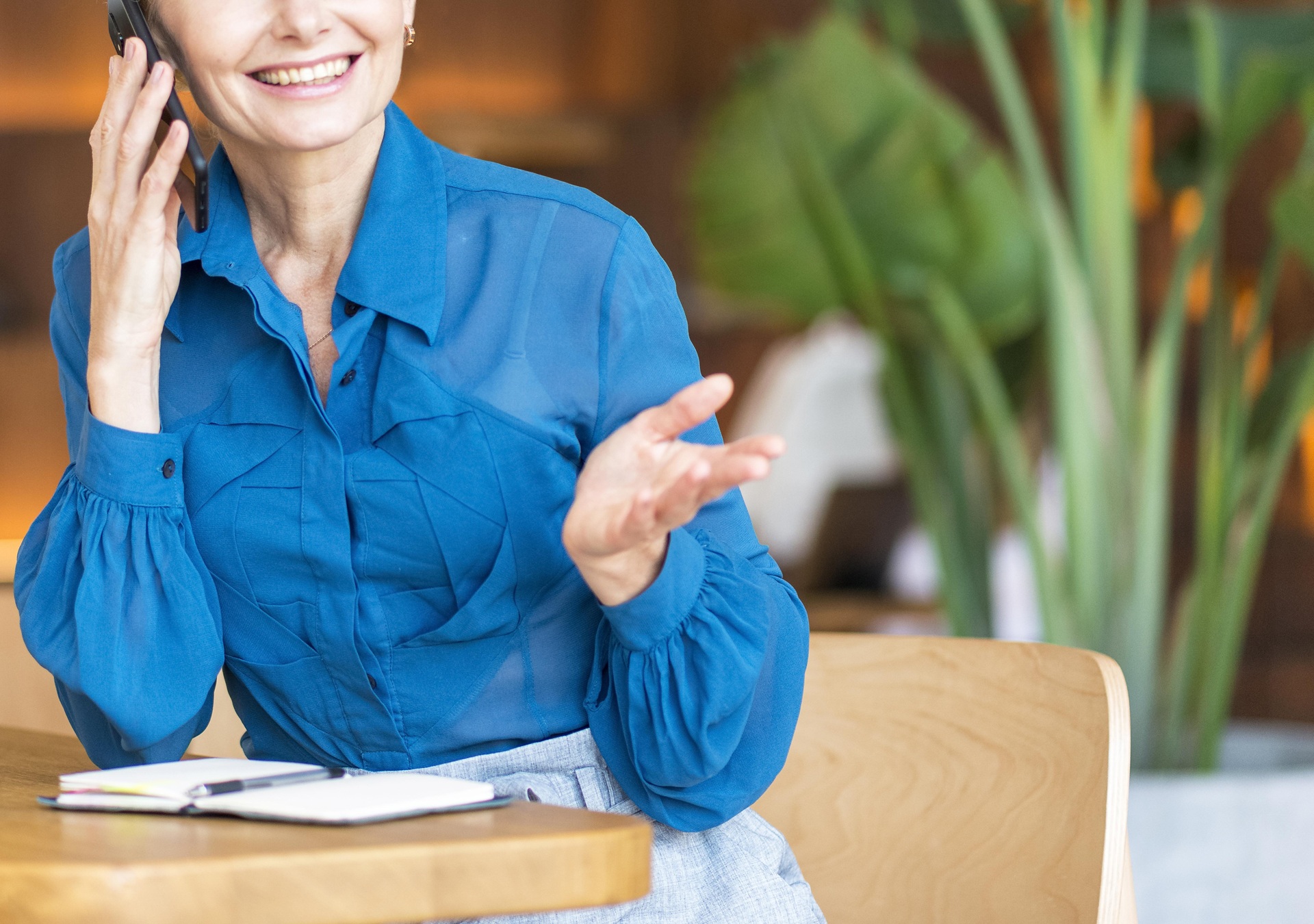A woman in a blue blouse sits at a table with an open notebook, smiling and gesturing while talking, with green plants in the background. A woman in a blue blouse sits at a table with an open notebook, smiling and gesturing while talking, with green plants in the background.