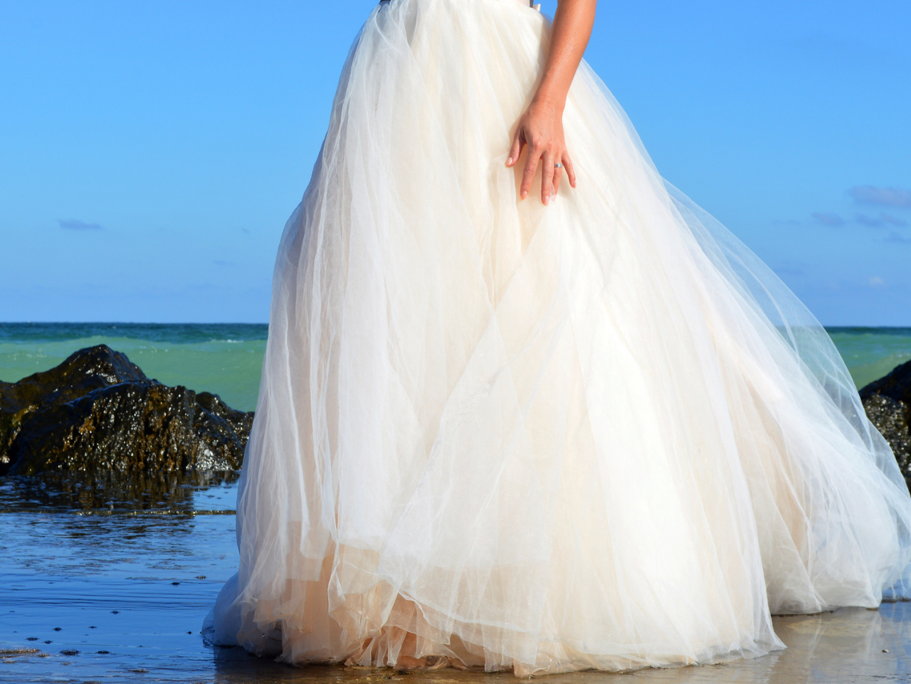 A person wearing a white wedding dress stands on a beach near rocks, with the ocean and blue sky in the background.