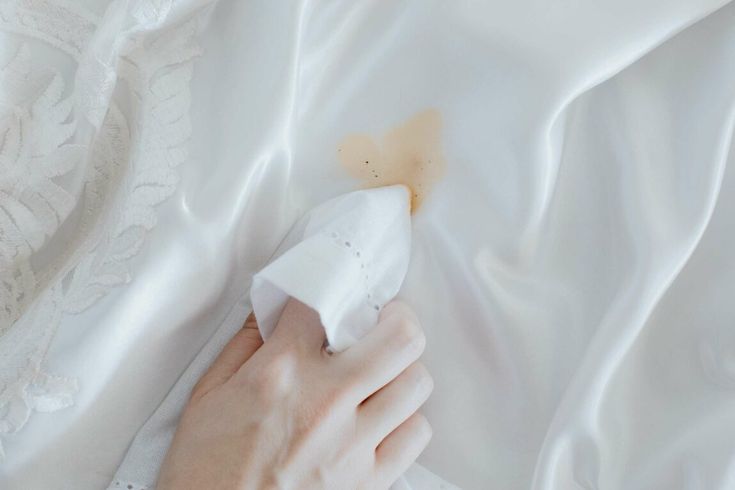 A hand uses a white cloth to blot a small brown stain on a white silk or satin fabric.