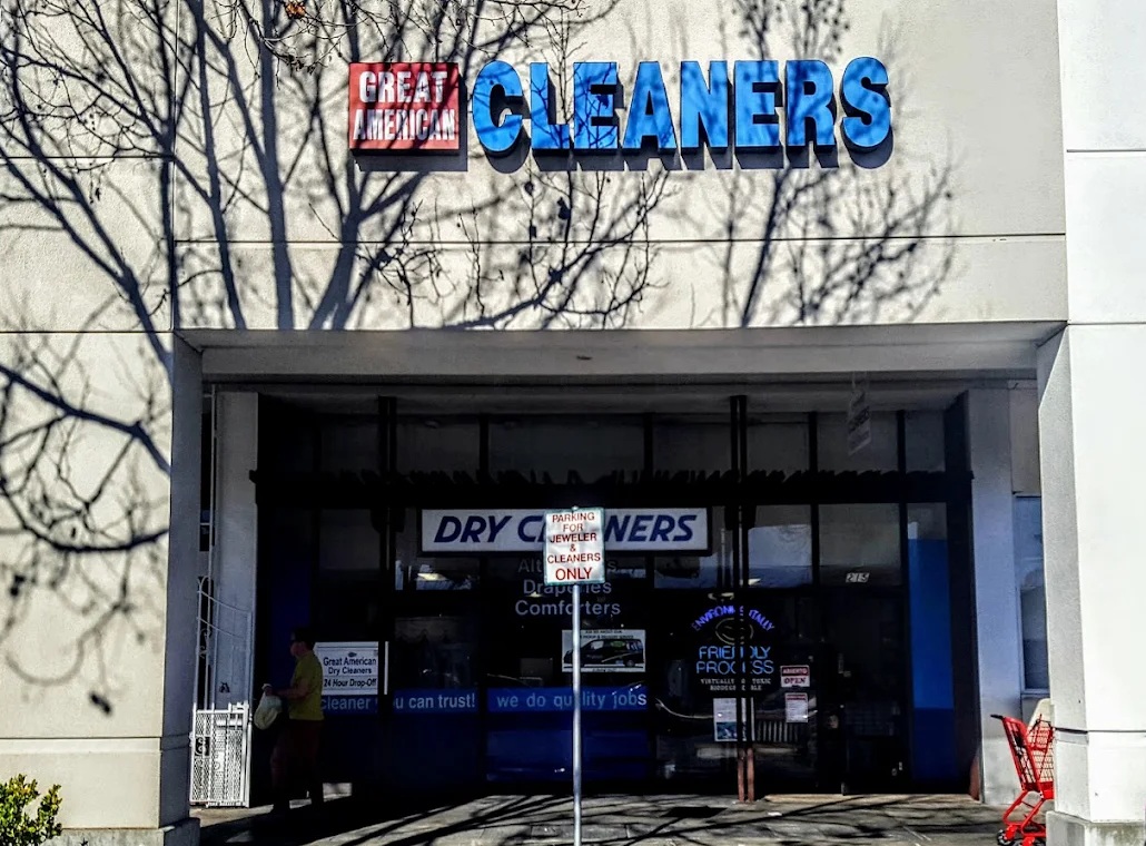 Storefront of "Great American Cleaners" with a blue and white sign, a shopping cart outside, and tree shadows cast on the building.
