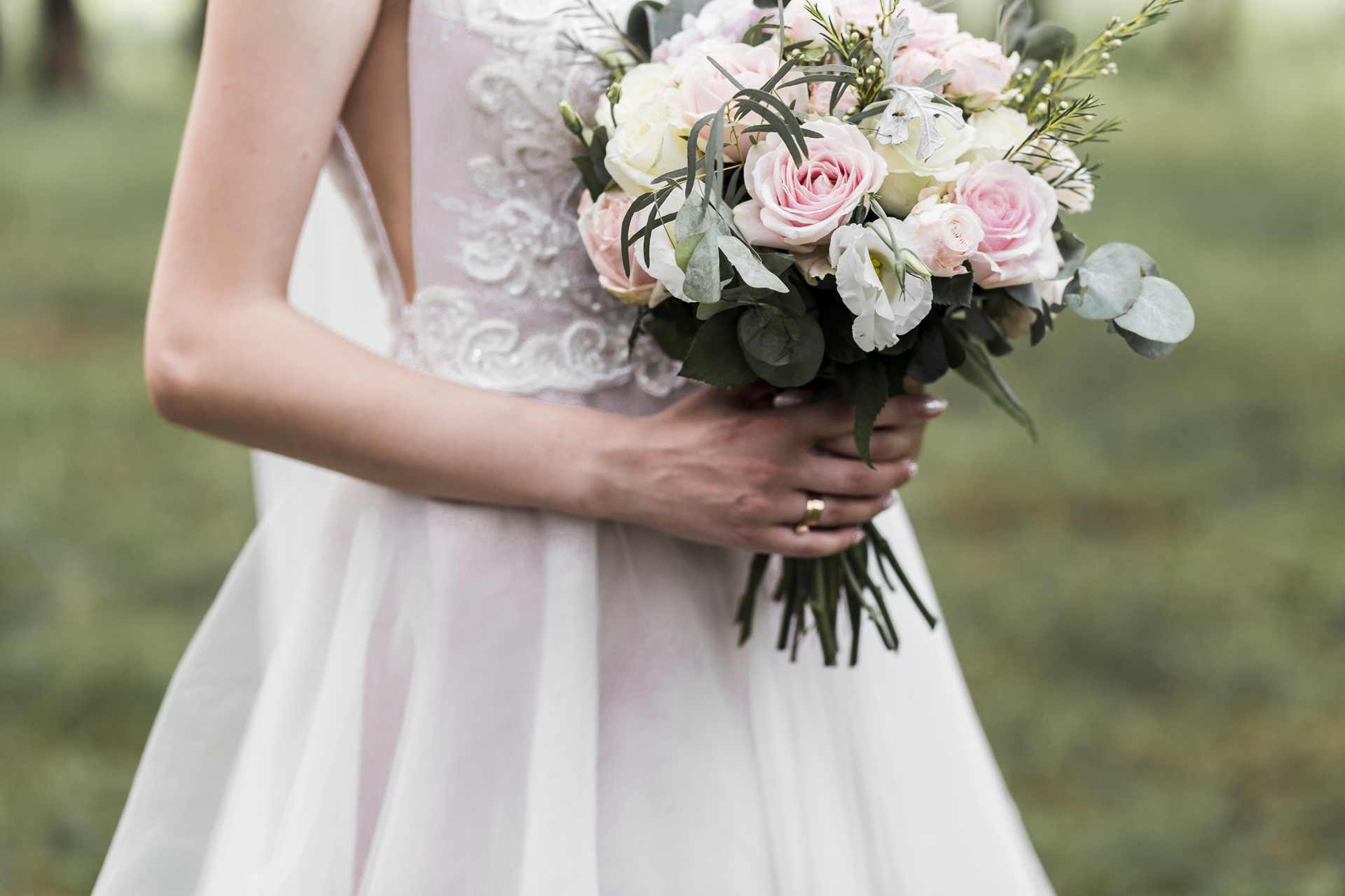 A bride in a white wedding dress holds a bouquet of pink and white flowers with greenery, standing outdoors.