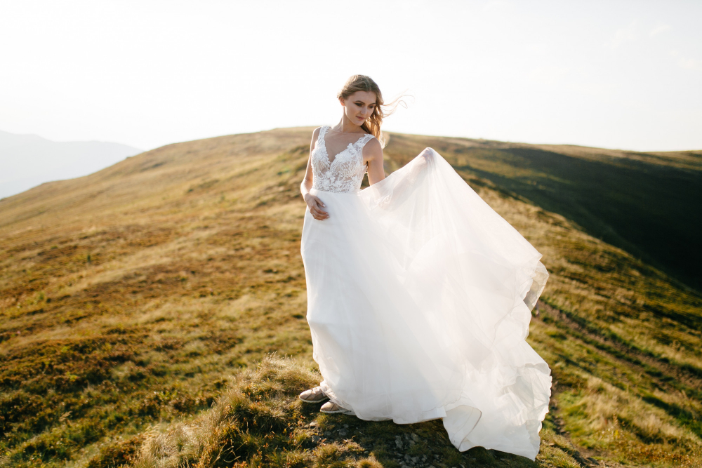 A woman in a white wedding dress stands on a grassy hilltop with wind blowing her dress, under a clear sky.