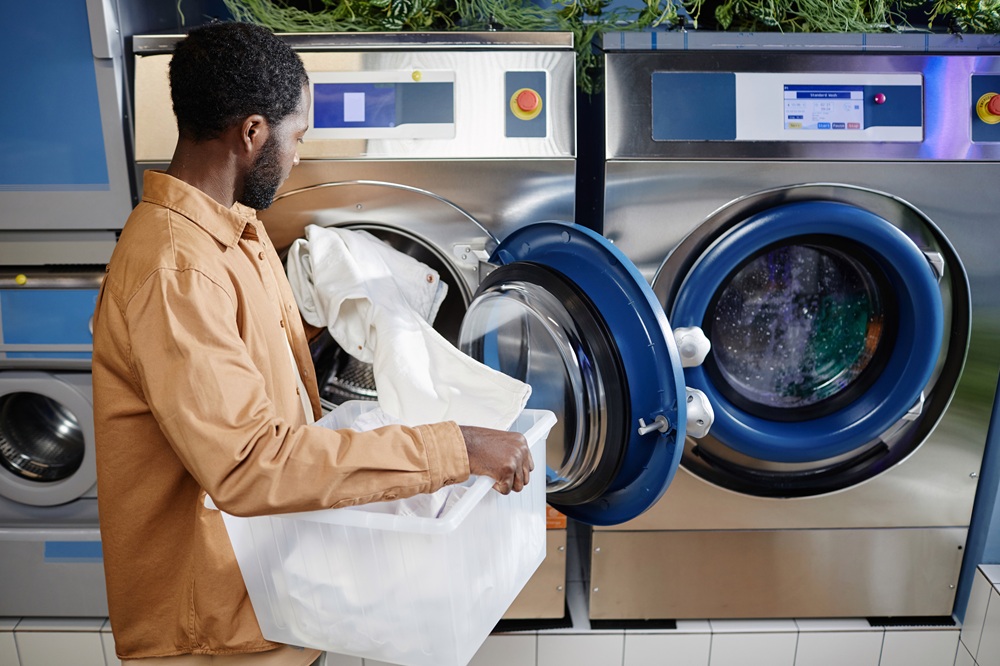 A man loads white laundry into a front-loading washing machine at a laundromat, holding a plastic basket.