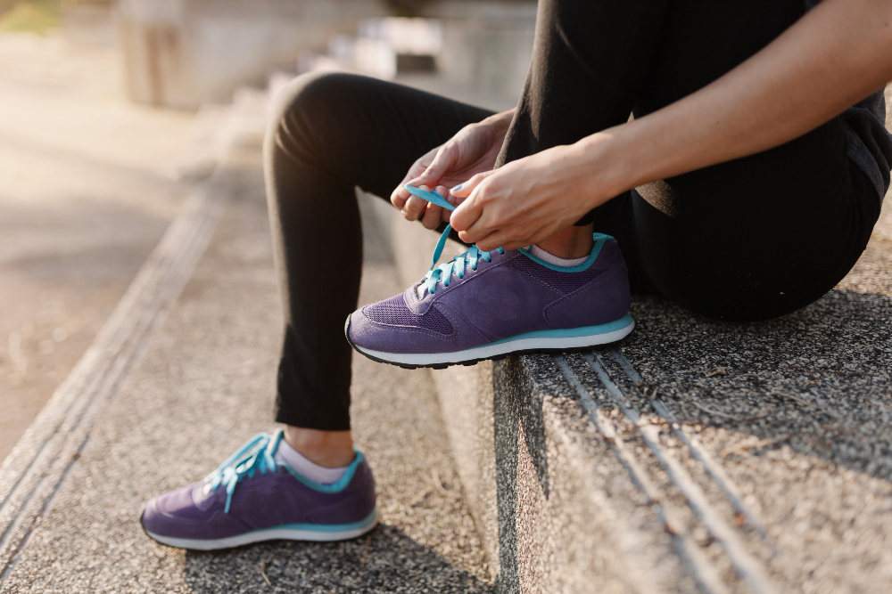 Person sitting on outdoor steps tying the laces of their purple athletic shoes, wearing black leggings—perfect for those in need of a professional shoe repair service in El Cerrito CA to keep their favorite sneakers looking fresh.