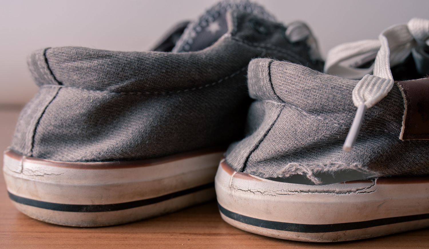 A close-up of two worn gray canvas sneakers on a wooden surface, showing frayed fabric and cracked soles.