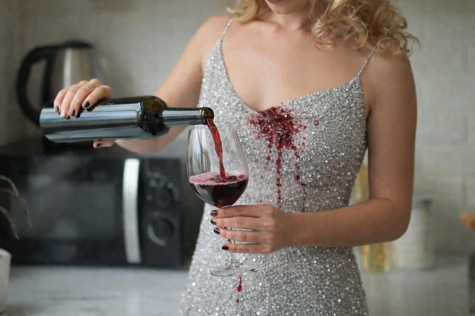 A woman in a sparkly dress spills red wine on her chest while pouring it into a glass in a kitchen setting.
