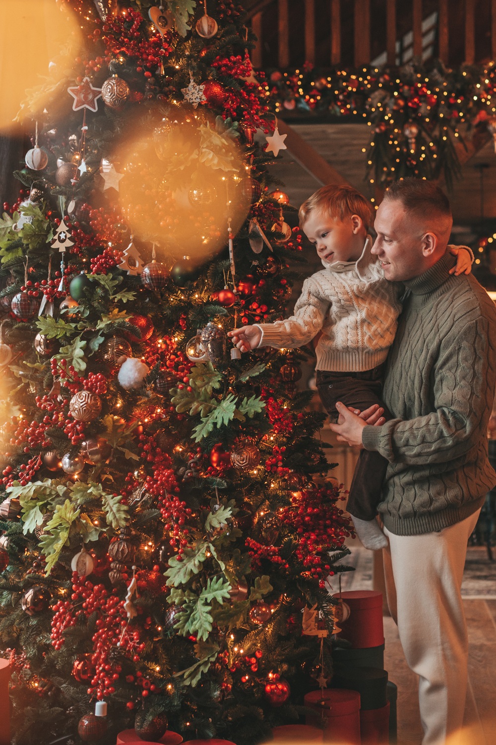 An adult holds a child next to a decorated Christmas tree with ornaments, red berries, and lights. Wrapped gifts are placed under the tree.
