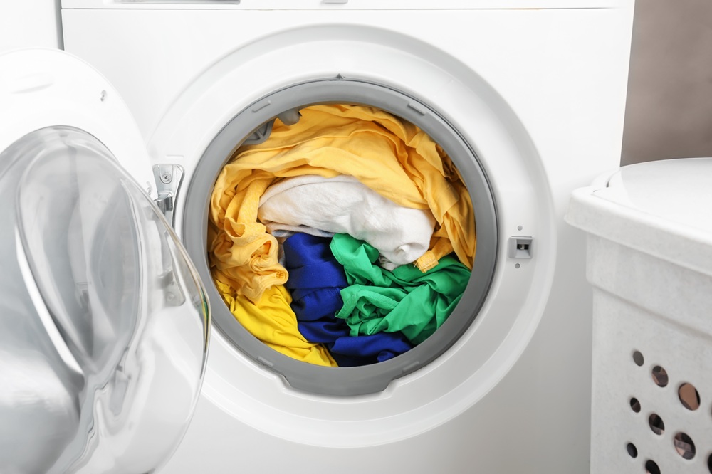 Front-loading washing machine with its door open, filled with yellow, white, blue, and green clothes next to a laundry basket. Front-loading washing machine with its door open, filled with yellow, white, blue, and green clothes next to a laundry basket.