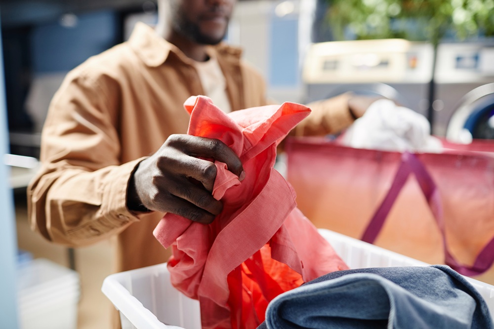A person sorting laundry, holding a red shirt over a basket with other clothes in a laundromat.