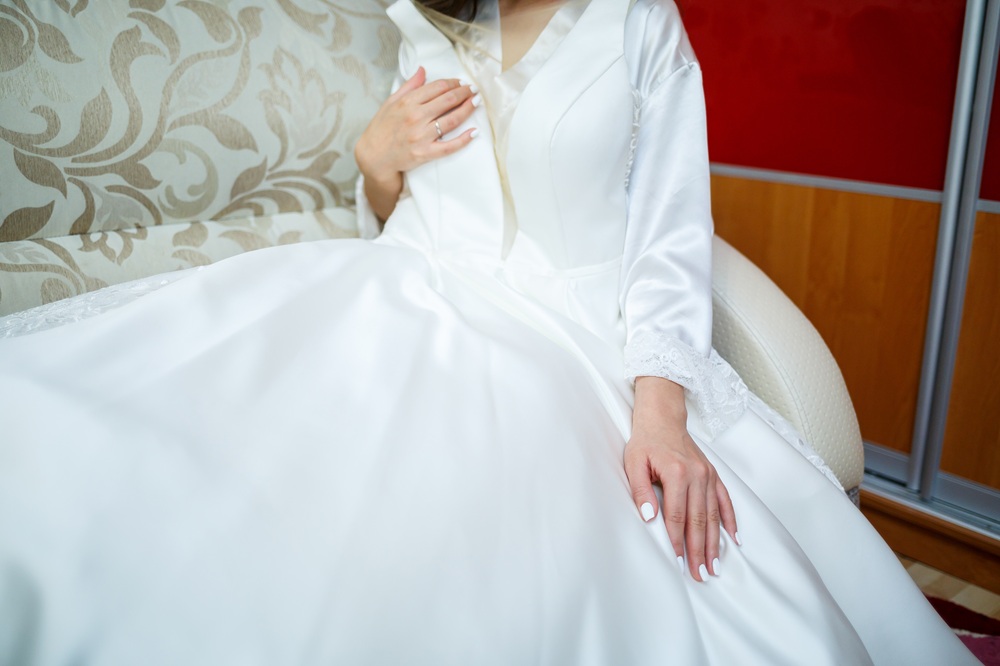 A person in a white satin wedding dress sits on a patterned sofa, with hands resting on their lap and chest, showing a close-up of the gown and manicured nails.