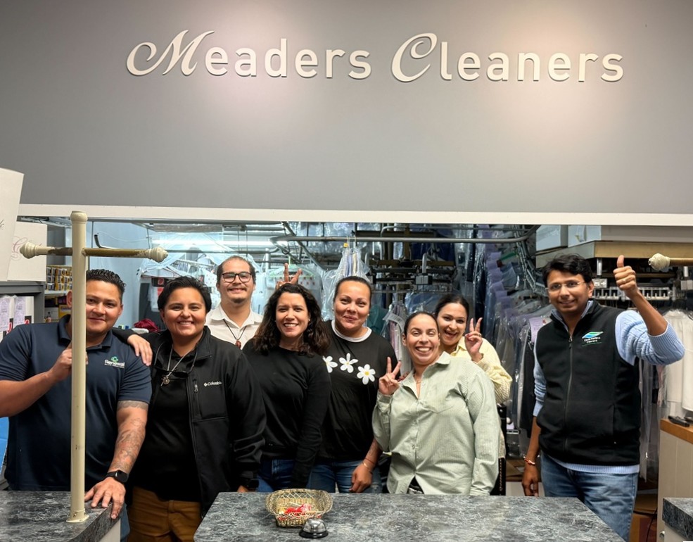 Eight people stand and smile behind the counter at Meaders Cleaners, posing for a group photo inside the dry cleaning shop.