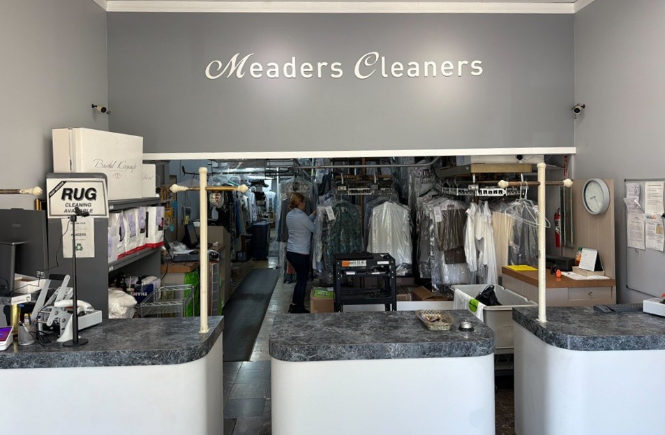 Front counter of a dry cleaning business with clothing racks, equipment, and staff visible in the workspace behind the counter. "Meaders Cleaners" sign is displayed above.