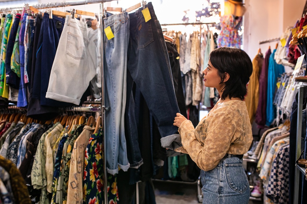A woman browses through a rack of jeans and shirts in a clothing store, holding up a pair of dark jeans for a closer look.