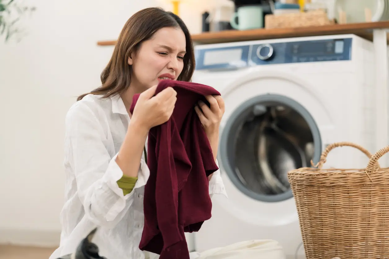 A woman with a disgusted expression smells a maroon garment in front of a washing machine and laundry basket.